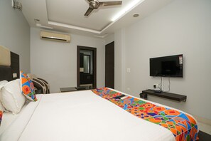 Una habitación de hotel con cama, ventilador de techo, equipo de aire acondicionado, televisor y pequeño escritorio con silla.