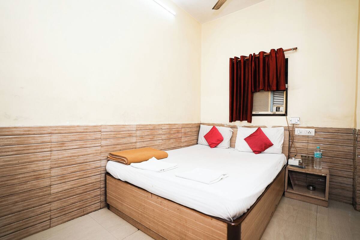 A neatly made bed with a brown headboard, a small bedside table with a water bottle, and a window with red curtains.