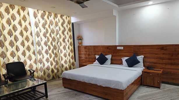 A bedroom with a wooden headboard, a glass coffee table, a bed with white bedding and two pillows, a nightstand, and a patterned curtain. - Hotel Grand (New Delhi)