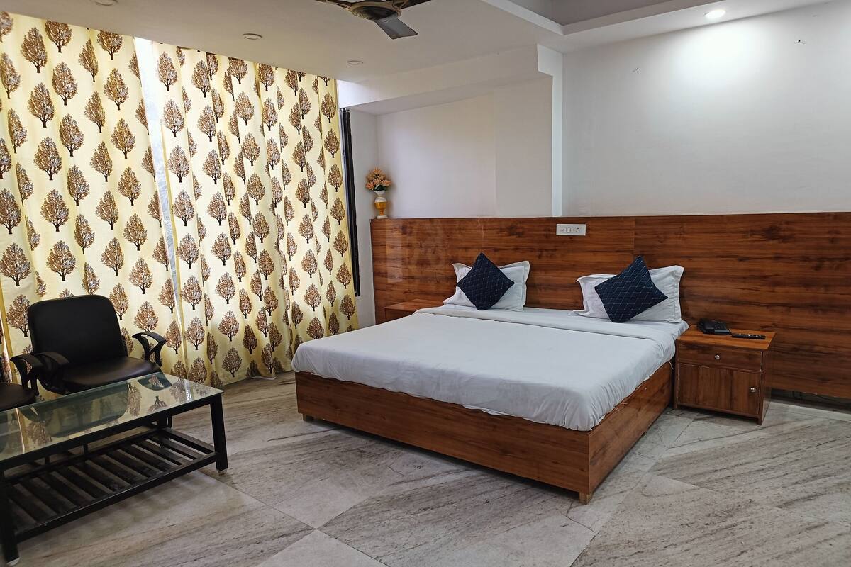 A bedroom with a wooden headboard, a glass coffee table, a bed with white bedding and two pillows, a nightstand, and a patterned curtain.