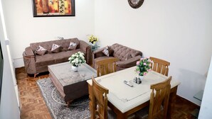 A living room with two brown sofas, a coffee table, and a dining table with chairs. There is a clock on the wall and a framed artwork above the sofa. - Greenleaf Apartment And Suites, Chittaranjan Park (New Delhi)