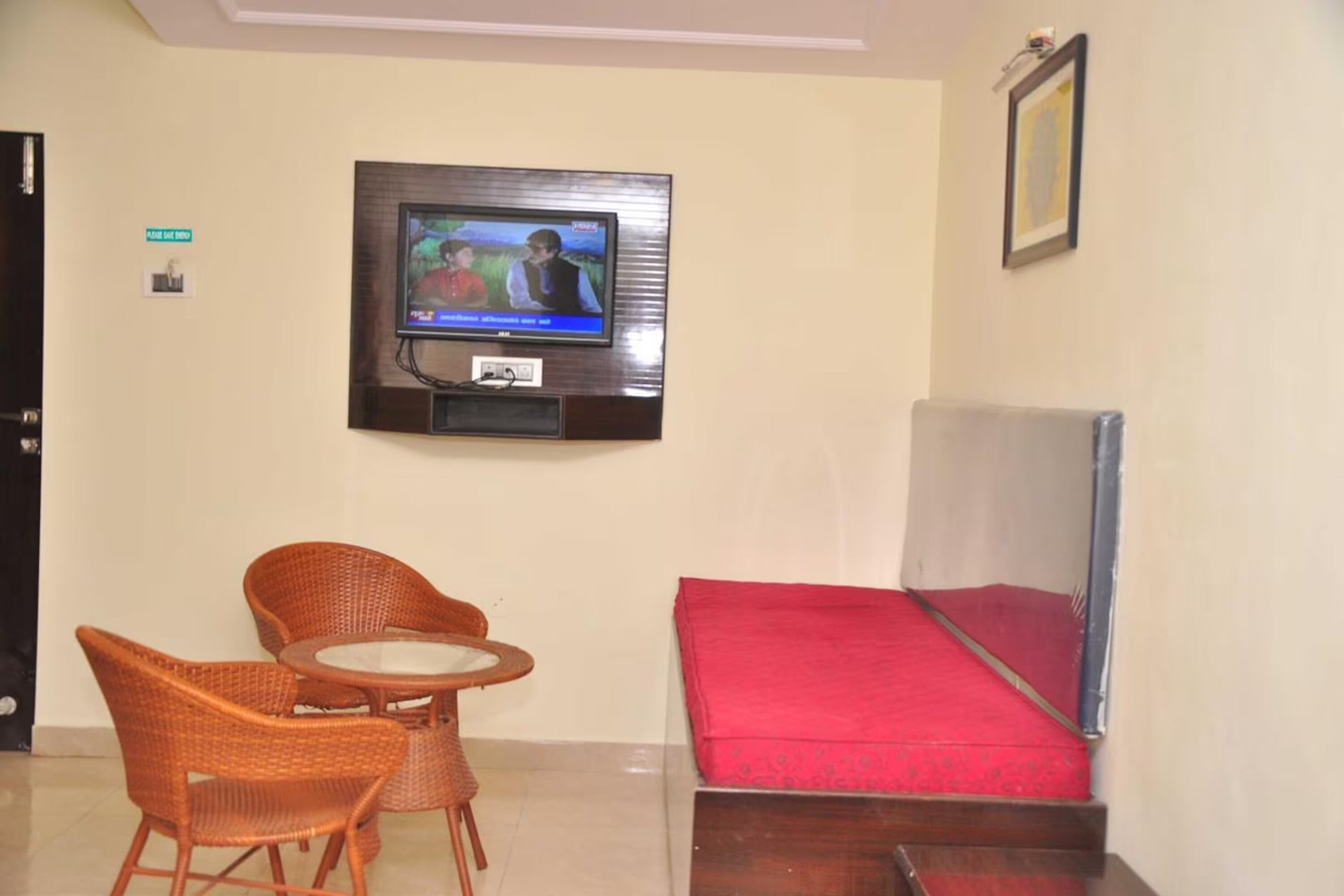 Quarto de hotel com televisão na parede, duas cadeiras de vime, uma mesa pequena e uma cama vermelha.