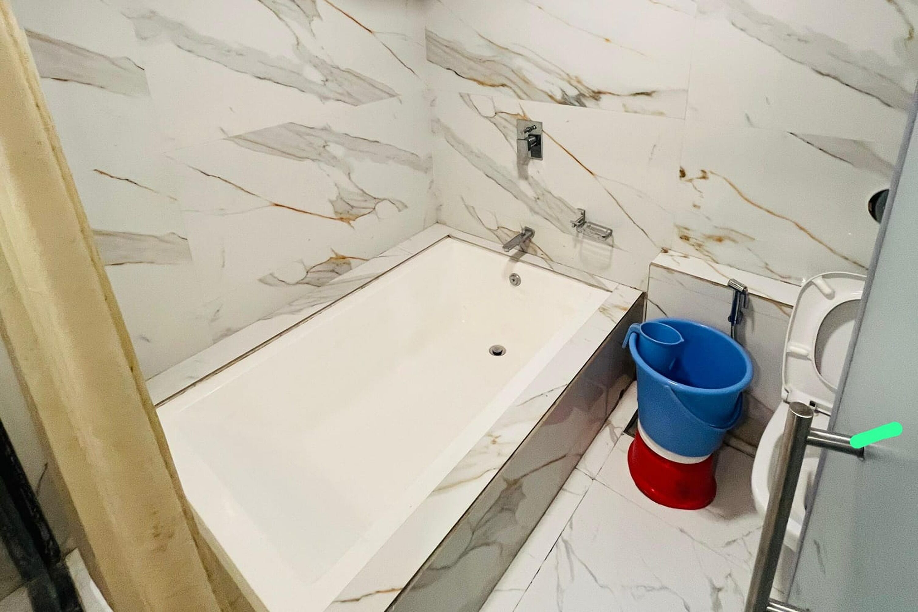 A bathroom with a marble wall, a bathtub, a toilet, and a blue and red bucket.
