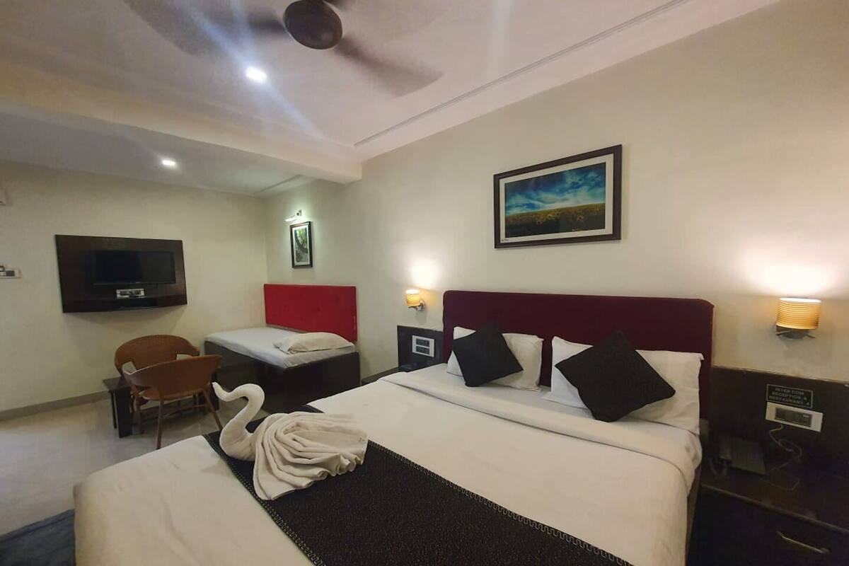Ein Hotelzimmer mit einem großen Bett, einem kleineren Bett, einem Fernseher, einem Sessel, einem Schreibtisch und einem Bild an der Wand.