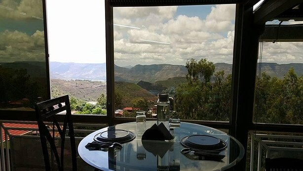 A dining table with a glass top, set with plates, glasses, and cutlery, overlooking a mountainous landscape through large windows. - Silver Leef (Wai)