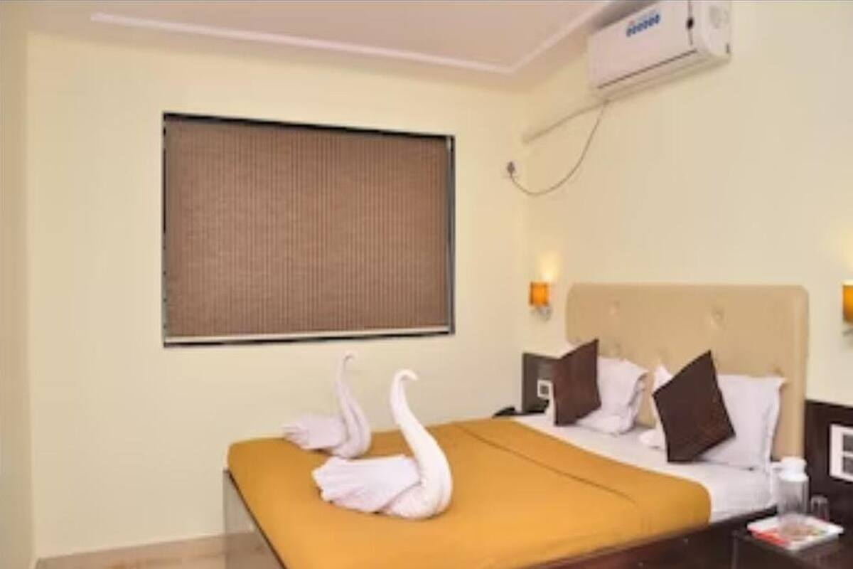 Ein Hotelzimmer mit einem Bett, zwei schwanenförmigen Handtüchern, einem Fenster mit Jalousien, einer Wand-Klimaanlage und einem Nachttisch mit einer Lampe.
