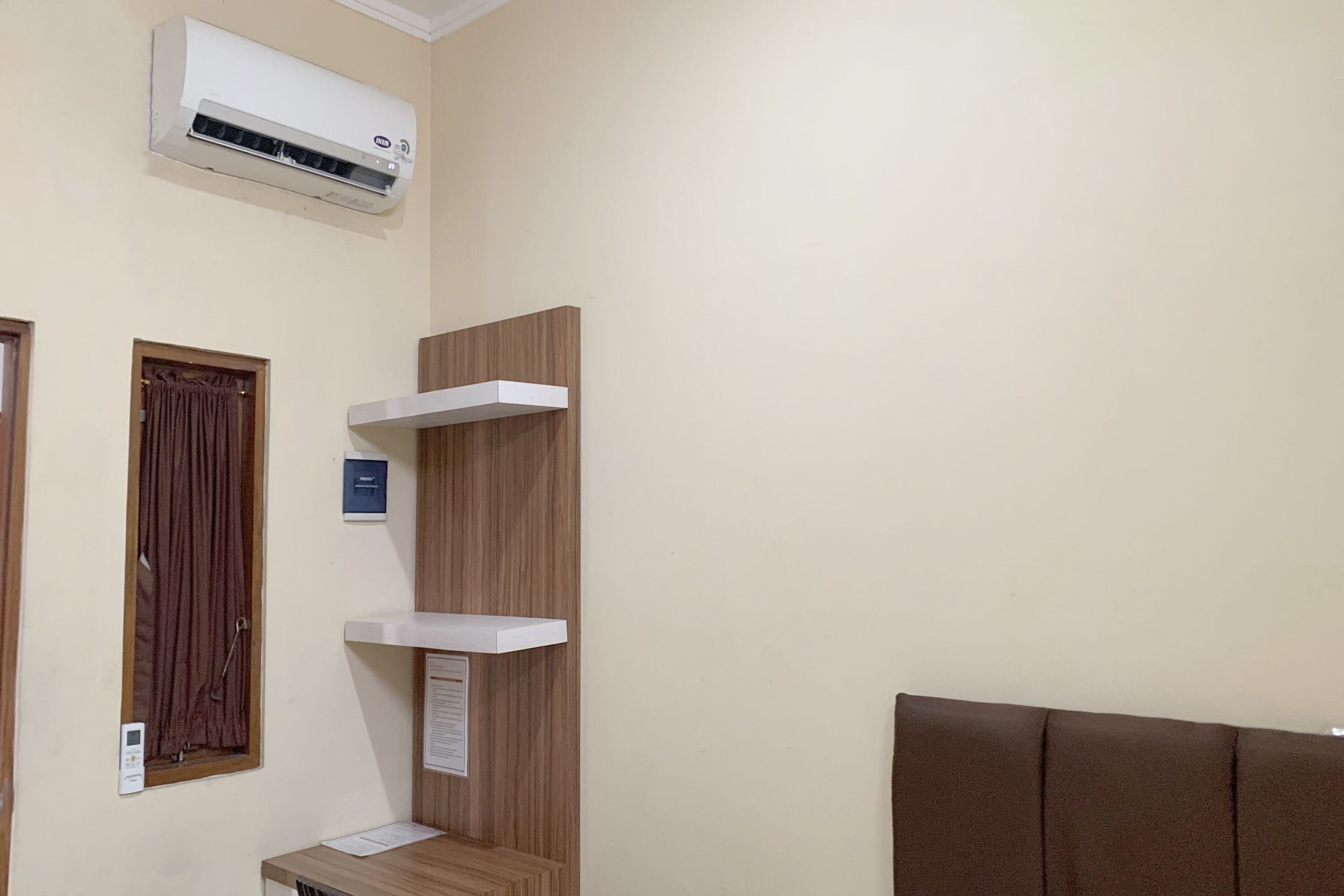 Une chambre d’hôtel avec une tête de lit en bois, un climatiseur blanc, une étagère en bois et une porte avec un rideau.