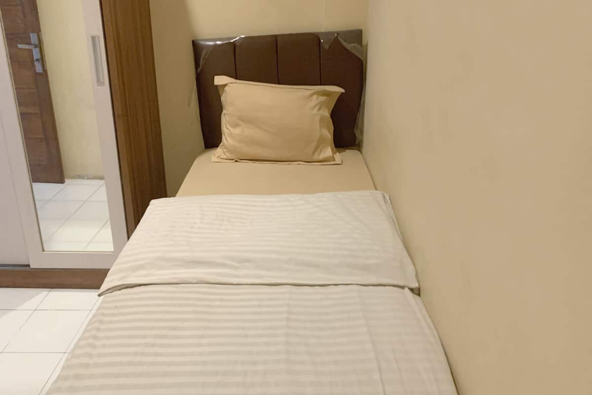 A small, neatly made bed with a headboard and a pillow in a room with plain walls and a tiled floor.