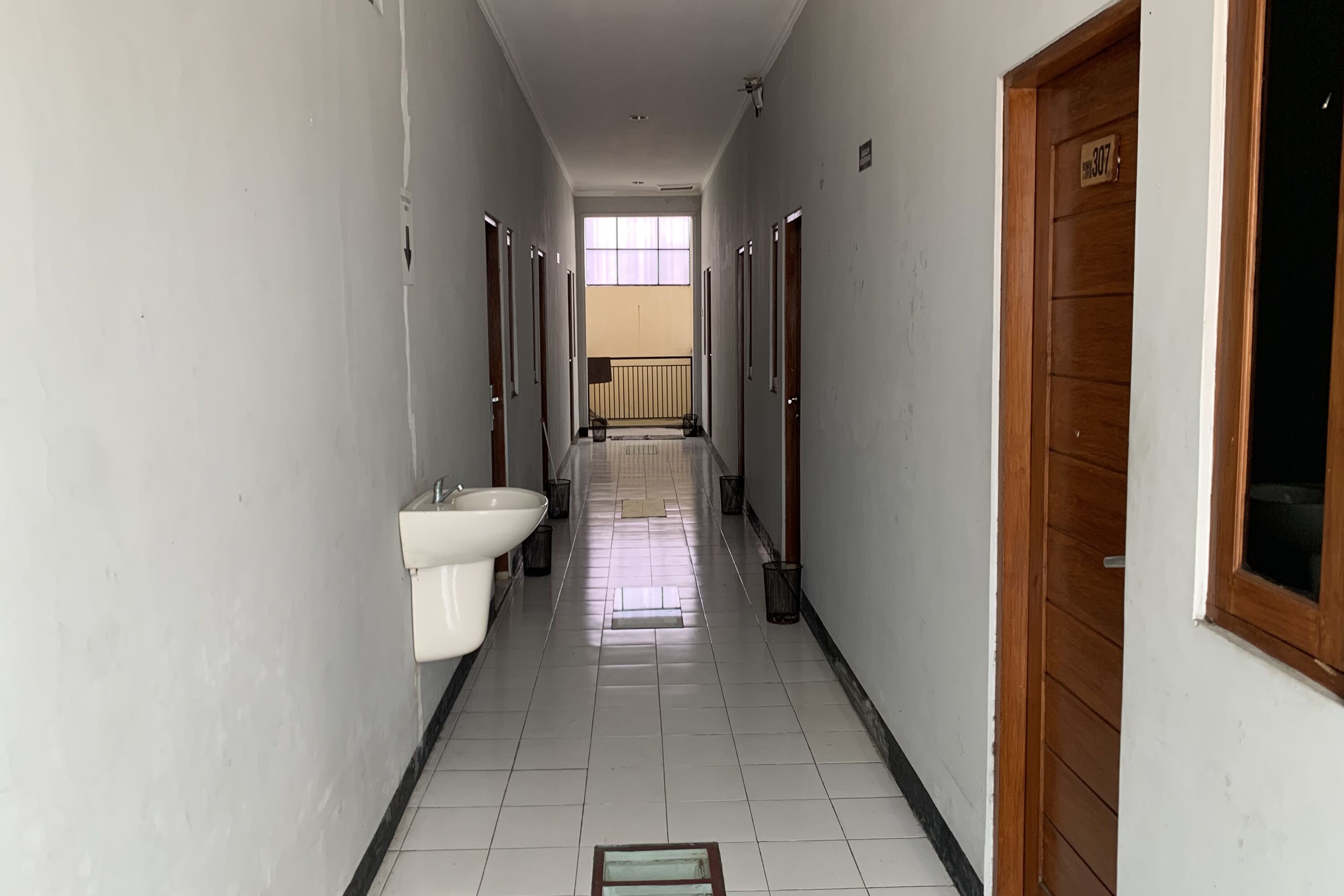 A corridor with a sink and bidets on one side, a wooden door on the other, and a window at the end.