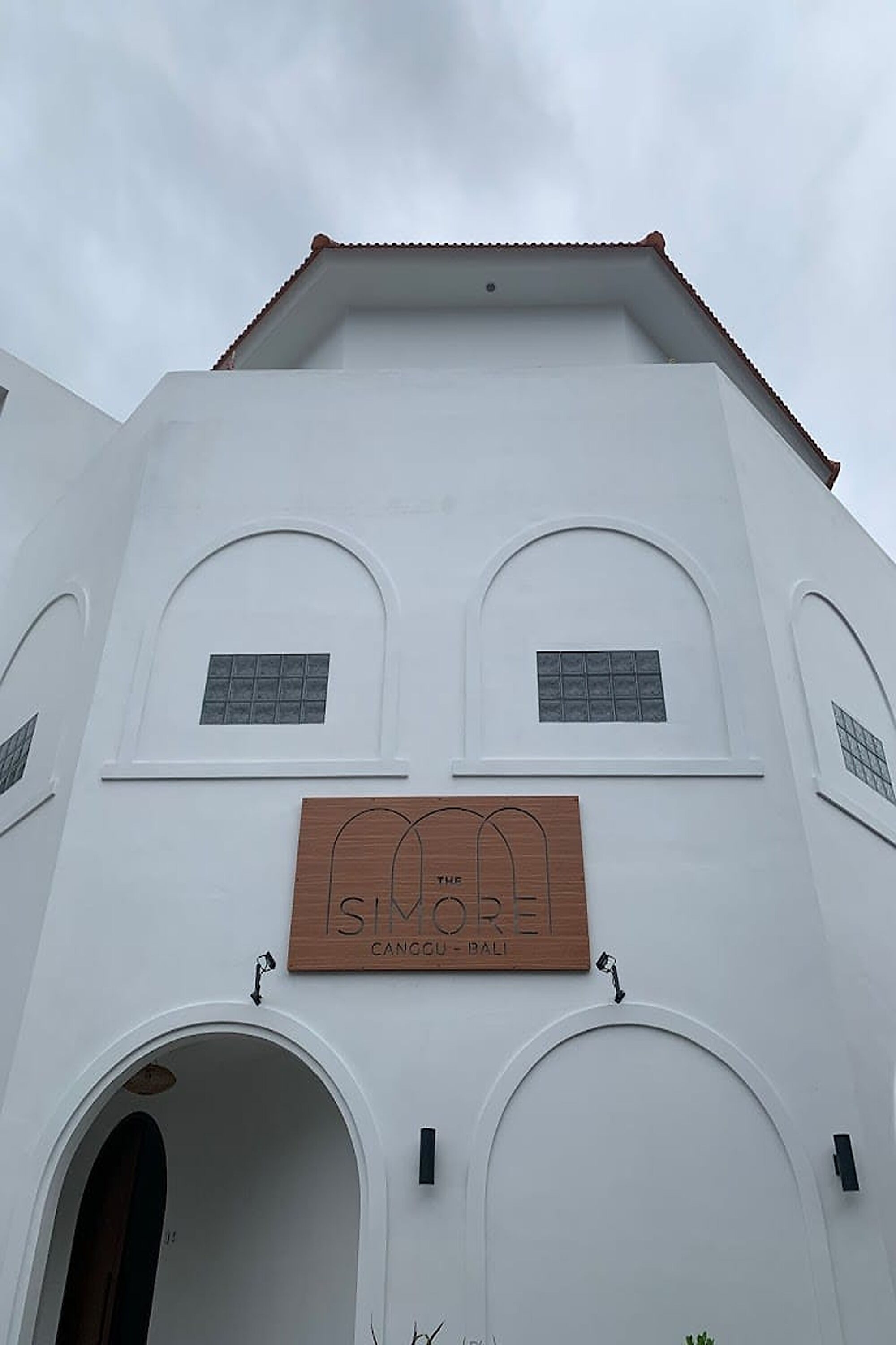 A building with a white facade, two arched windows, and a wooden sign that reads "SIMOREI Canggu Bali".