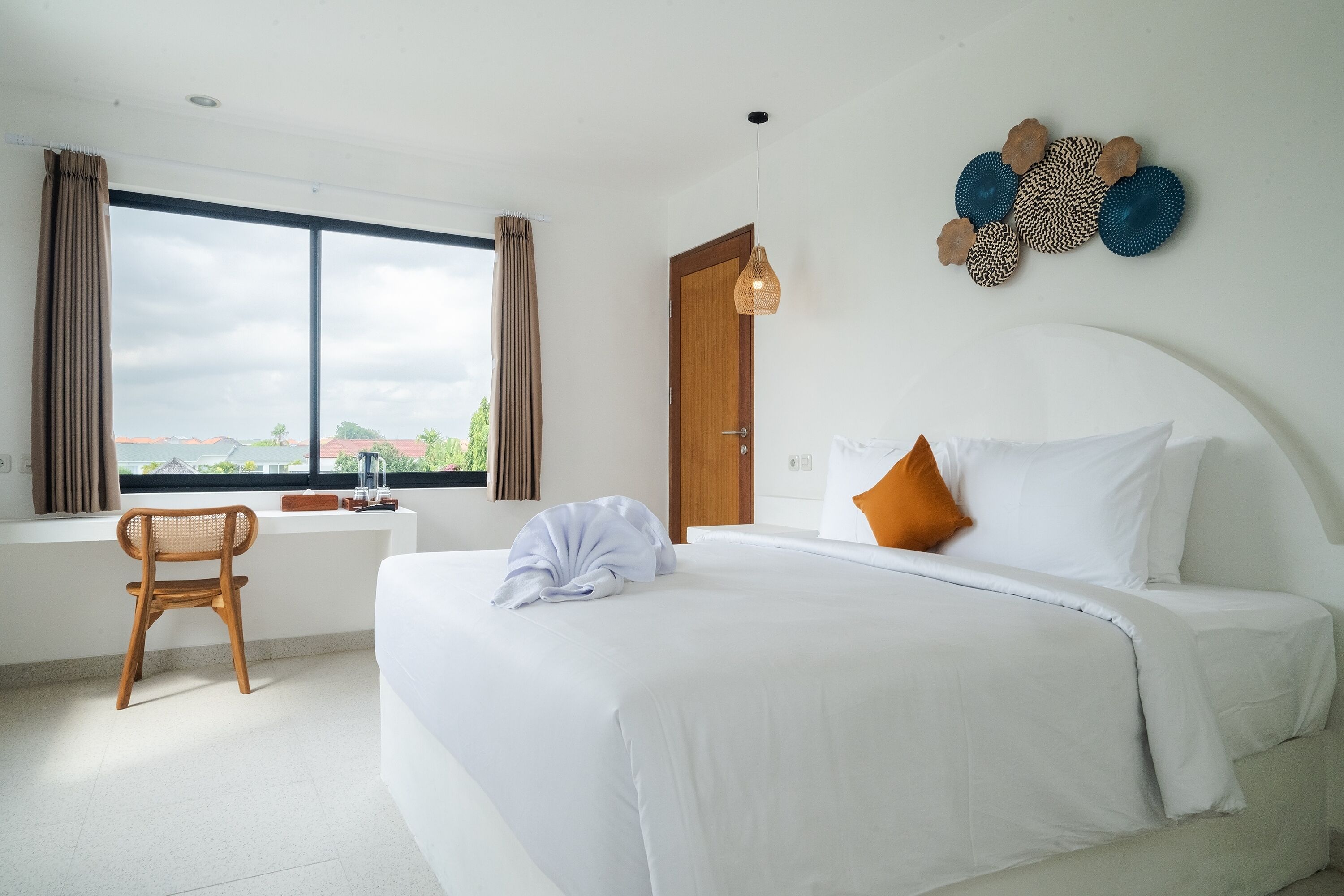 A hotel room with a large bed, a desk, and a chair. There is a window with curtains, a door, and wall art.