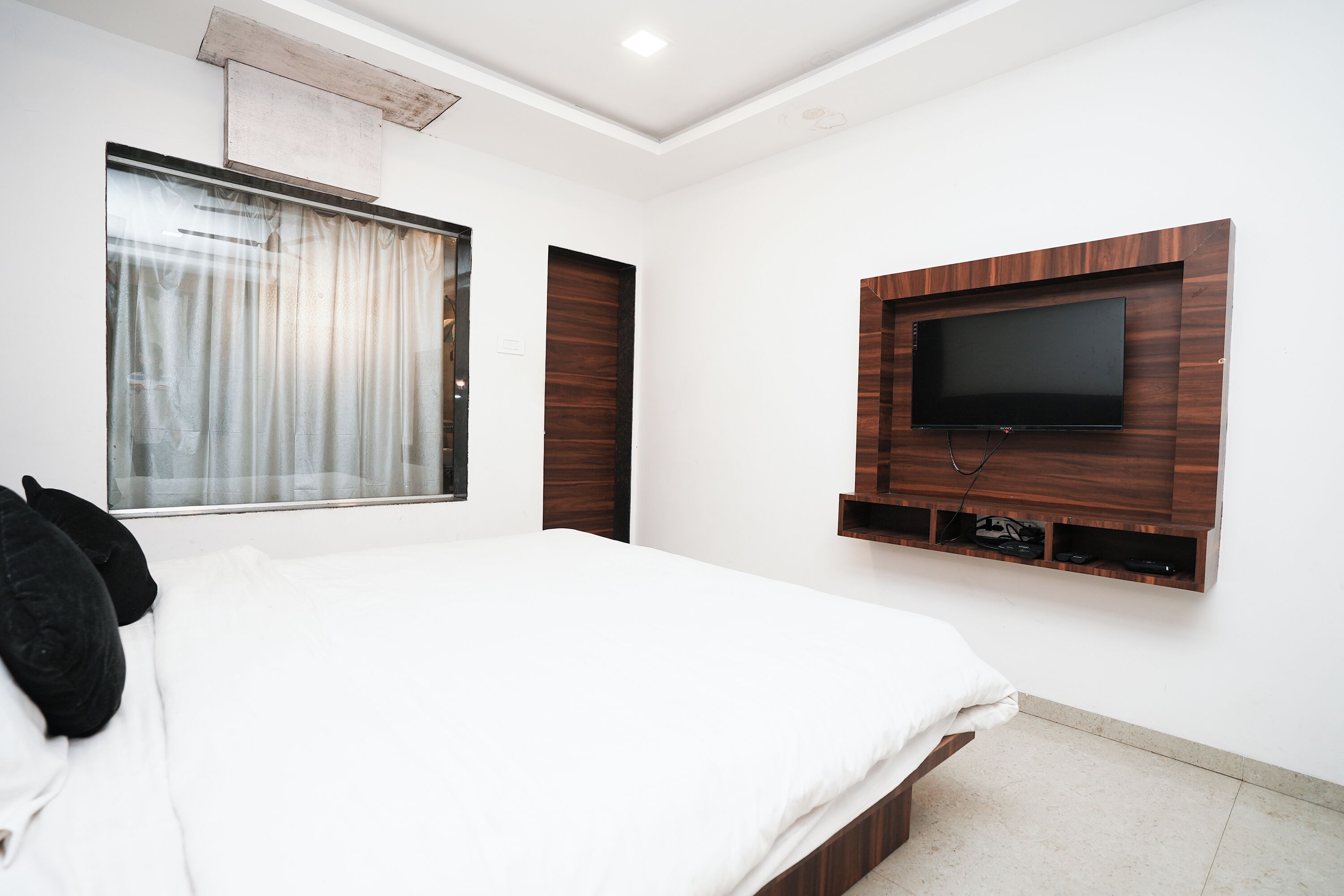 A bedroom with a large bed, a TV mounted on the wall, and a window with curtains.