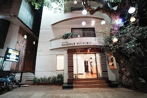 La façade de l’hôtel Maharaja Residency, avec une enseigne au-dessus de l’entrée, un escalier menant à la porte et un arbre décoré de guirlandes lumineuses.