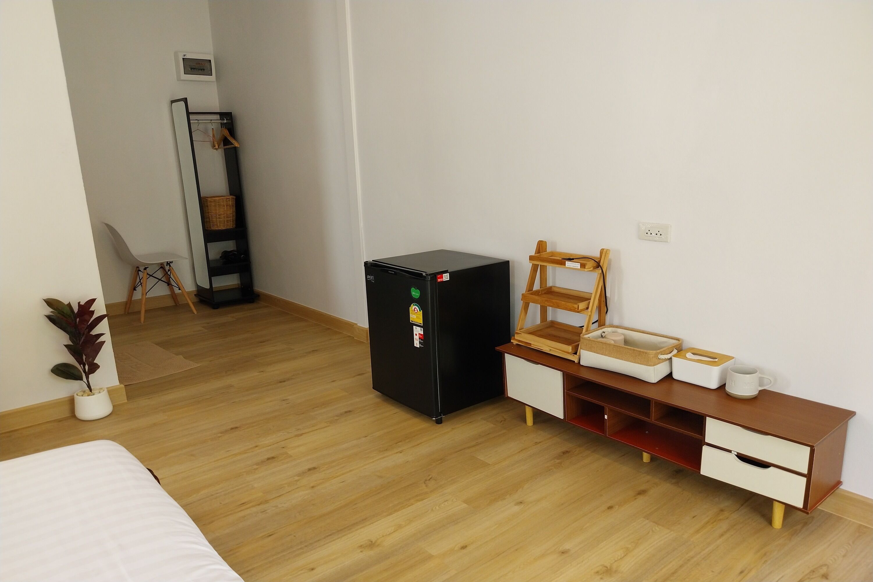 Una habitación compacta con cama, refrigerador negro, mesita de madera con cajones, una silla y un pequeño estante.