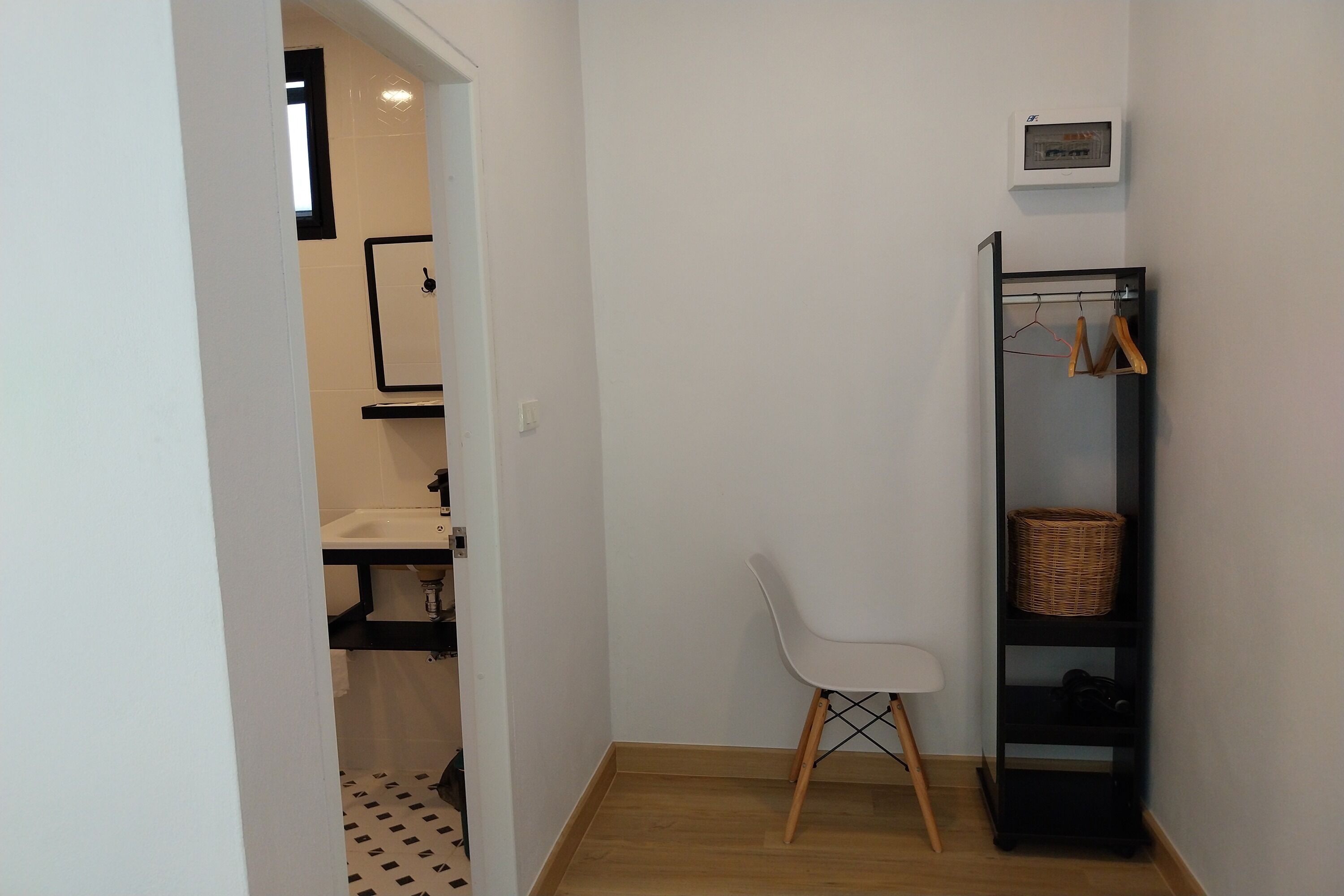 Un baño pequeño y moderno con un lavamanos blanco, un espejo y zona de ducha. Hay una silla blanca y un estante negro con una cesta.