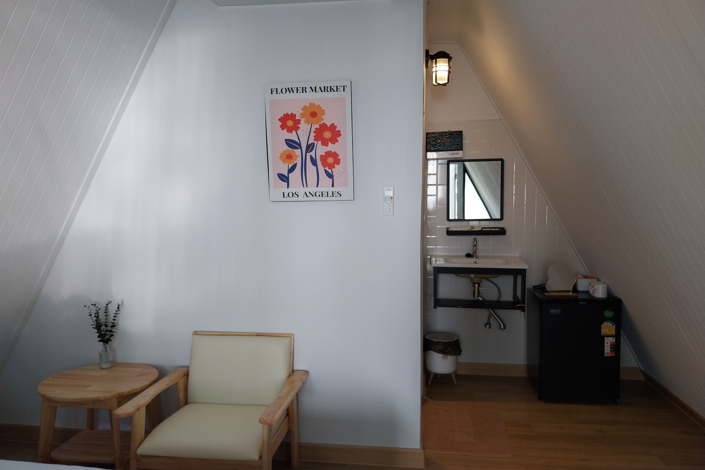 A small, cozy room with a chair, a small table, a wall-mounted picture, a sink, and a mirror.