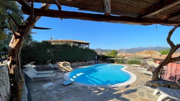 A swimming pool with stone flooring and lounge chairs, surrounded by a stone wall and a wooden pergola. - VILLA ELENA PRIVATE POOL (Sardegna)