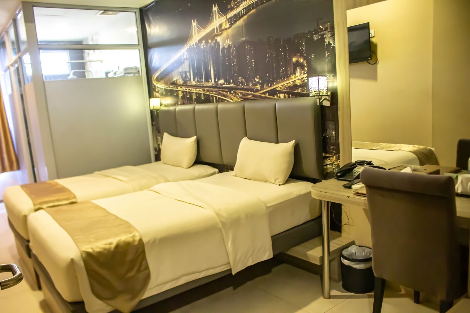 A hotel room with a bed, a desk, a chair, and a cityscape mural on the wall.