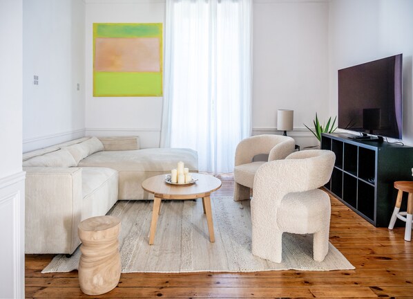 Una sala moderna con un sofá seccional, una mesa de centro redonda y dos sillones. Un televisor de pantalla plana está montado en la pared, y un ventanal con cortinas translúcidas deja entrar abundante luz natural.