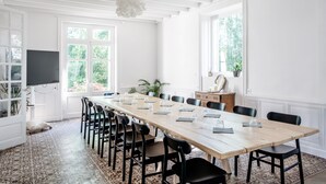 A long dining table with chairs, a television, and a potted plant in a bright room with large windows.