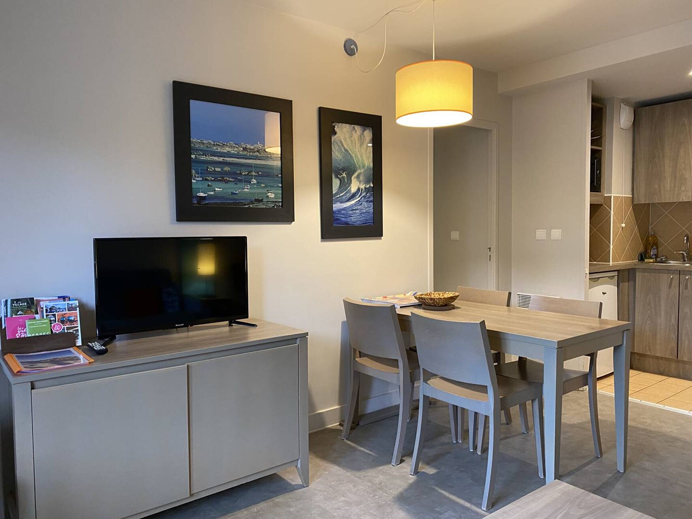 A modern living room with a flat-screen TV, dining table, and chairs. There is a kitchen area with wooden cabinets and a hanging light fixture.