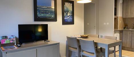 A modern living room with a flat-screen TV, dining table, and chairs. There is a kitchen area with wooden cabinets and a hanging light fixture.