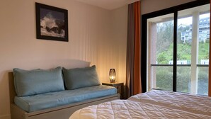 A bedroom with a bed, a sofa, a nightstand with a lamp, a window with a view of buildings, and a framed picture on the wall. - Stunning Beachfront Apartment with Pool, WiFi, and Balcony in Perros-Guirec (Perros-Guirec)