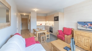 A modern, well-lit living room with a sofa, a coffee table, and a TV displaying a skiing scene. A kitchen with wooden cabinets and a dining area with a table and chairs are visible in the background. - 2-room apartment with parking in luxury residence in Alpe d'Huez (Huez)