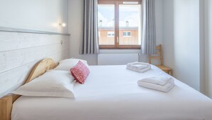 A neatly made bed with white linens and a red floral pillow, a wooden headboard, and a view of a building through a window with curtains. - 2-room apartment with parking in luxury residence in Alpe d'Huez (Huez)