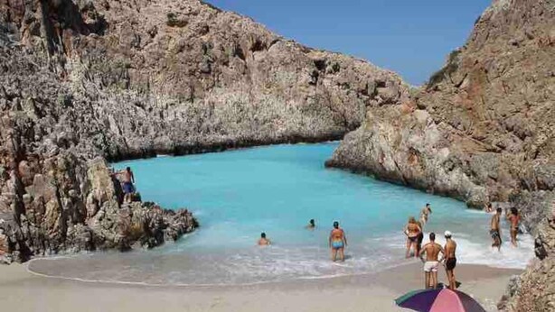 Ein felsiger Strand mit klarem türkisblauem Wasser, Menschen beim Schwimmen und Sonnenbaden, und eine Person mit einem Strandsonnenschirm.