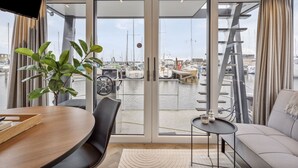 Ein modernes Zimmer mit einem großen Schiebeglasfenster, das zur Marina führt, einem Esstisch, einer Couch und einem kleinen Beistelltisch.