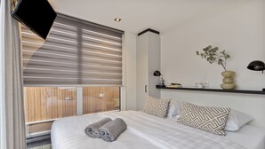 Ein modernes Hotelzimmer mit einem großen Bett, einem an der Wand befestigten Fernseher, einem Nachttisch mit Lampe und einer Zierpflanze.
