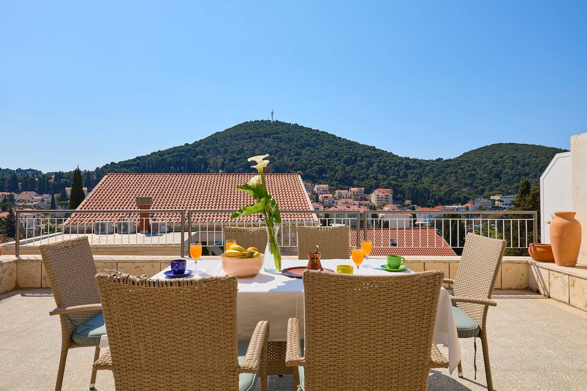 Um terraço com cadeiras de vime, uma mesa posta com uma tigela de frutas e vista para uma colina com edifícios de telhado vermelho.