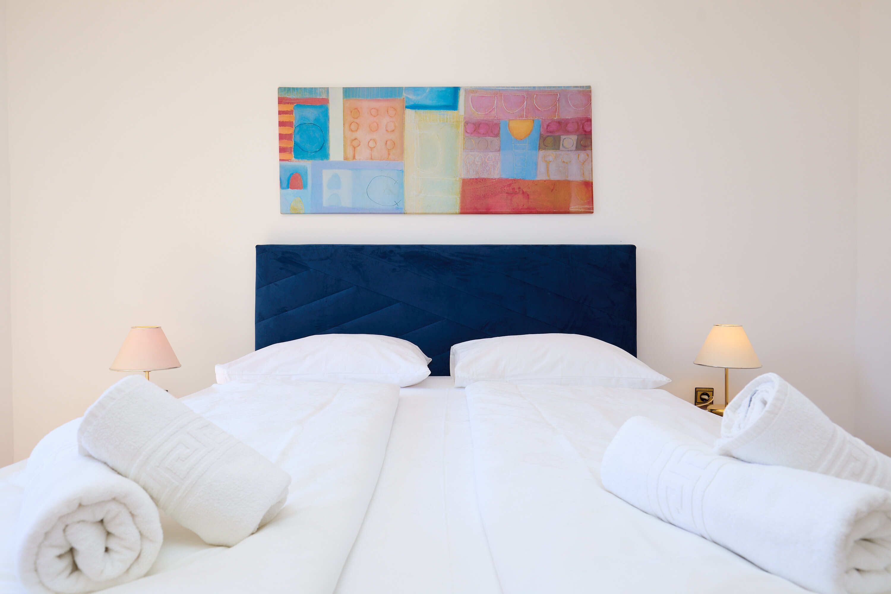 A neatly made bed with white linens and rolled towels, a blue headboard, and a colorful abstract painting above it.