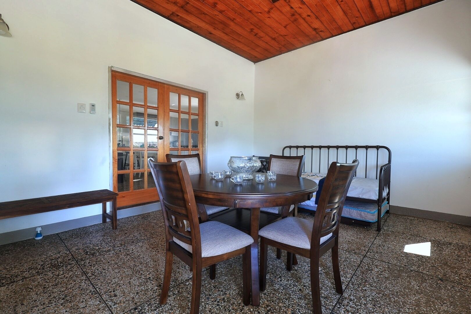 A dining area with a round wooden table, four chairs, a bench, and a metal bed frame against the wall.