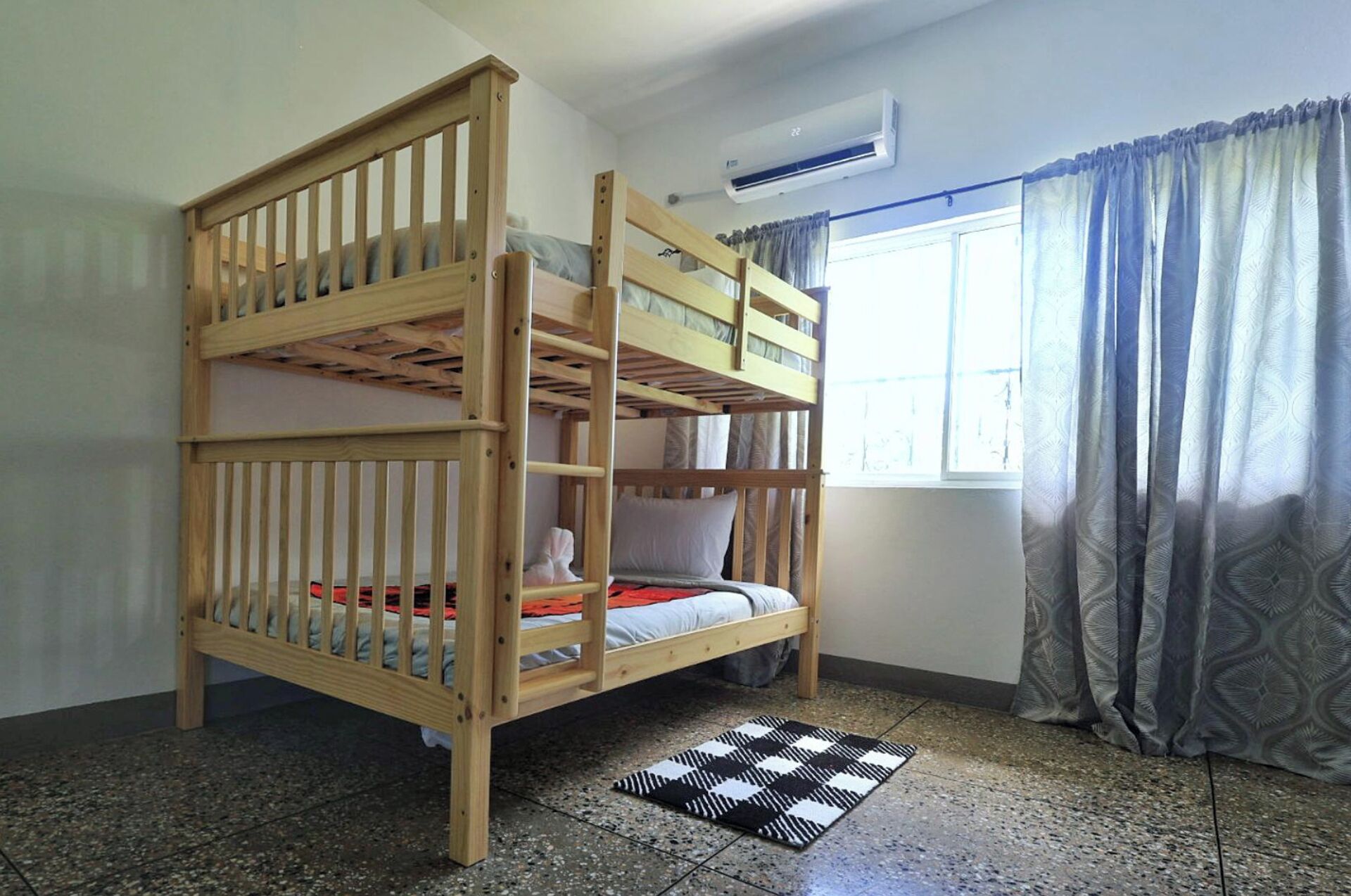 A bunk bed with a window and curtains, an air conditioner, and a patterned rug.