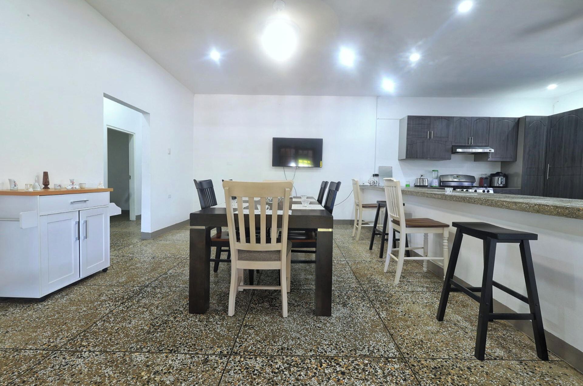 A modern kitchen with dark cabinets, a dining area with a table and chairs, and a small TV mounted on the wall.