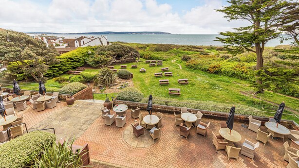An outdoor patio with wicker furniture and umbrellas, overlooking a grassy area with benches and a view of the sea. - The Beach House (Lymington)