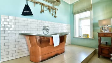 A bathroom with a copper bathtub, white tiled walls, a wooden shelf with a hairdryer, and a window with blinds.