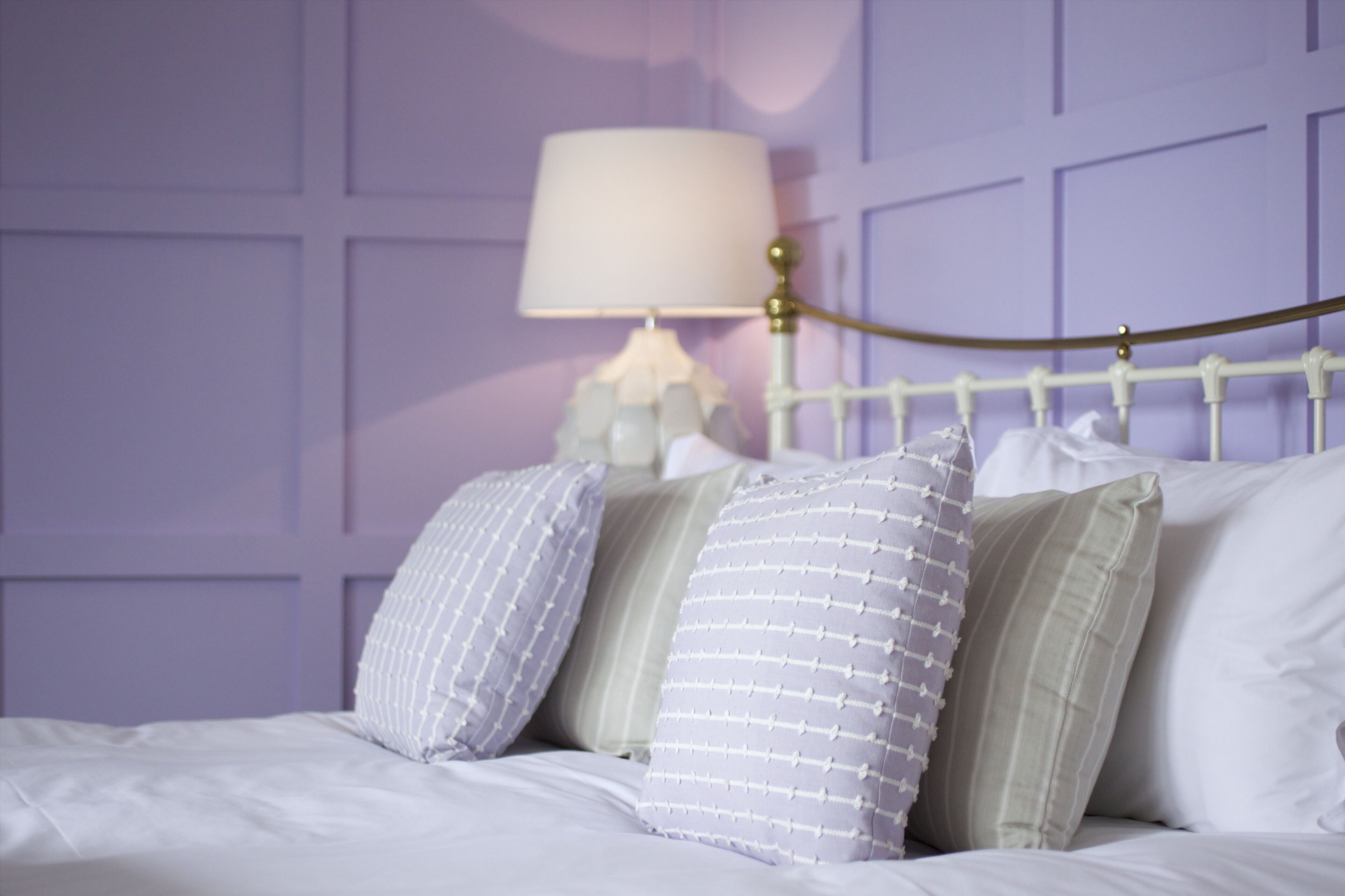 Un lit avec une literie blanche et de nombreux oreillers, une tête de lit en métal blanche et une lampe blanche avec un abat-jour beige, le tout sur un mur violet.