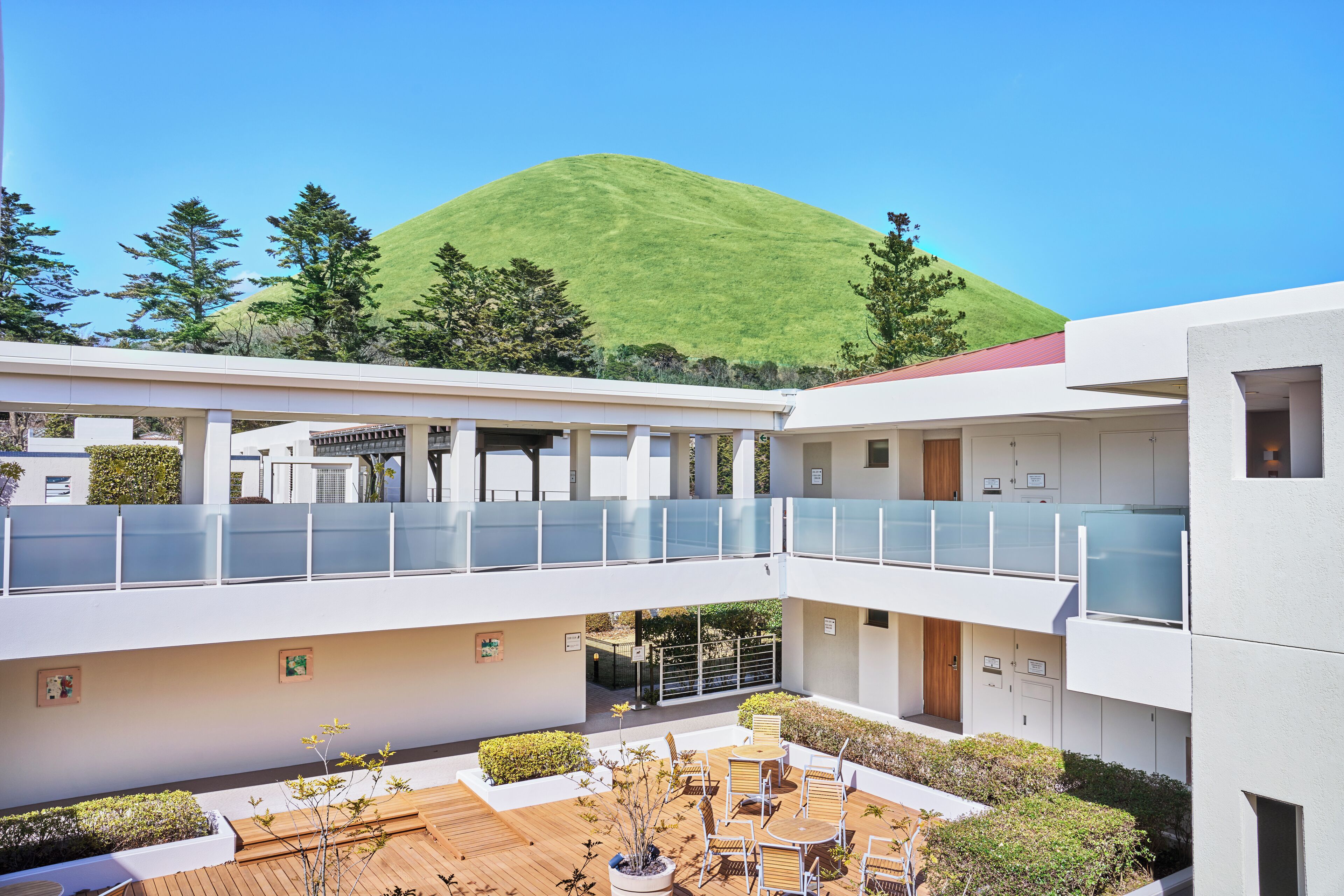 A modern building with a courtyard, outdoor seating, and a view of a green hill.