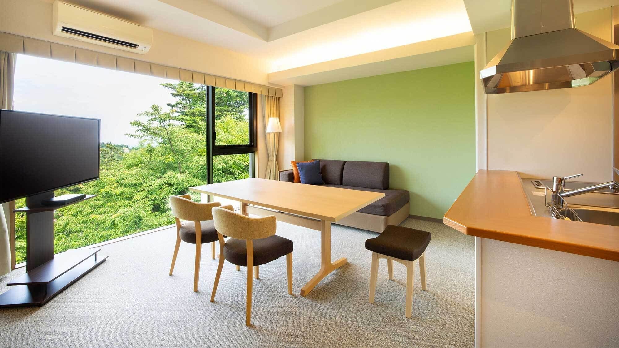 A hotel room with a dining table, chairs, a sofa, a TV, and a kitchen area with a sink.
