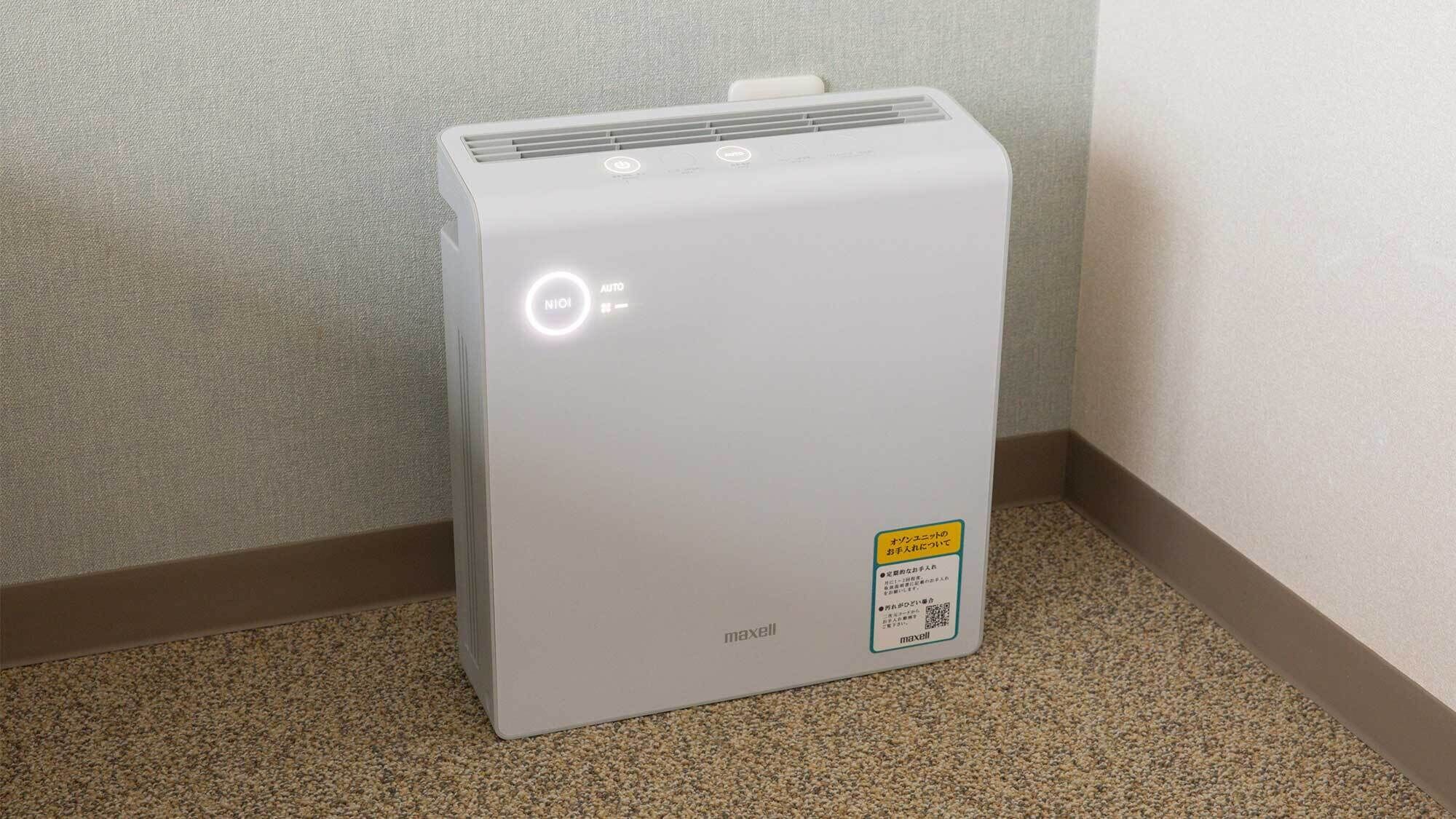 A white portable heater with a control panel and a warning label on the side, placed on a carpeted floor against a wall.