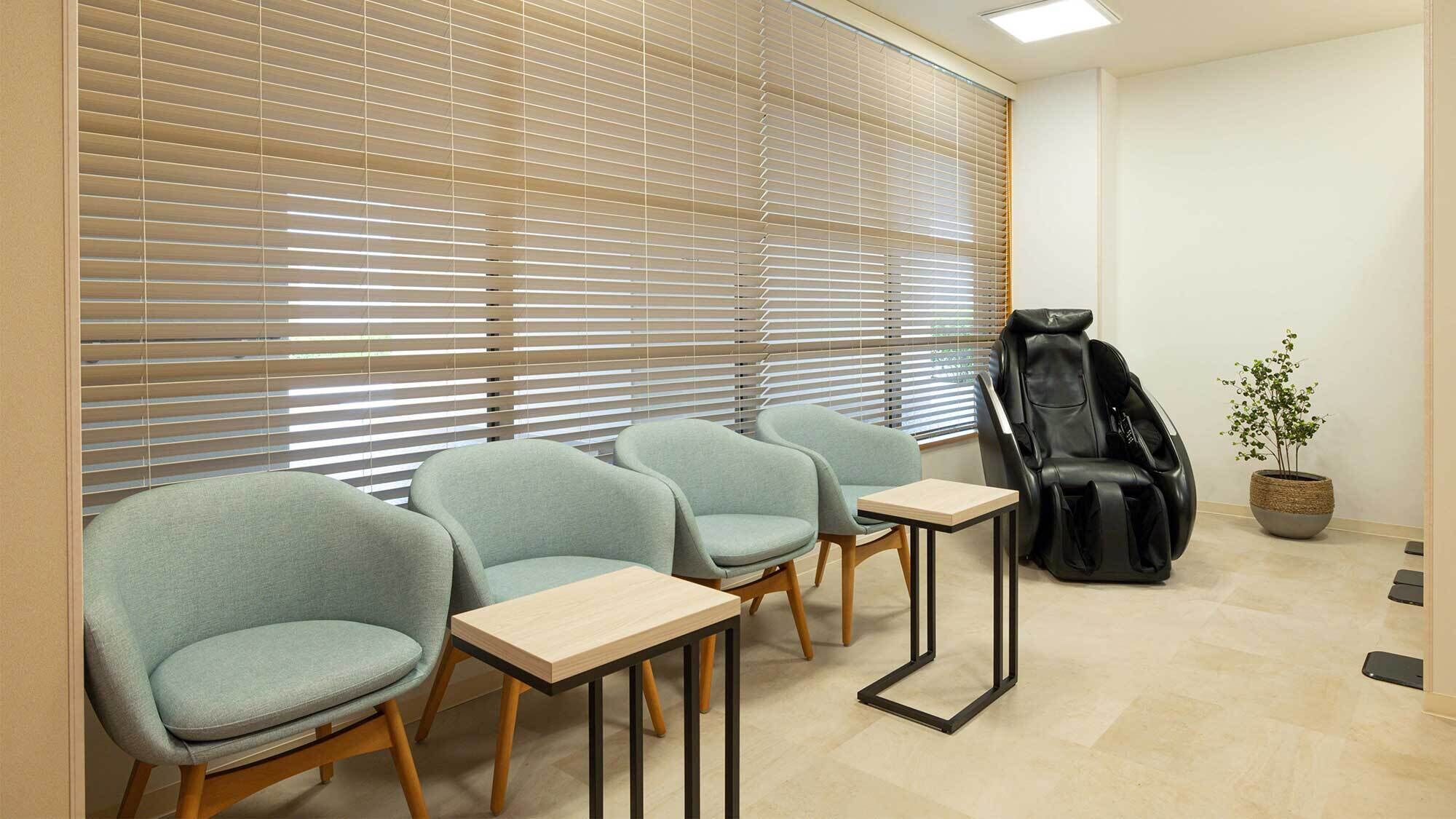 Une salle d’attente comprenant quatre chaises grises, un fauteuil de massage, une petite table et une plante en pot.
