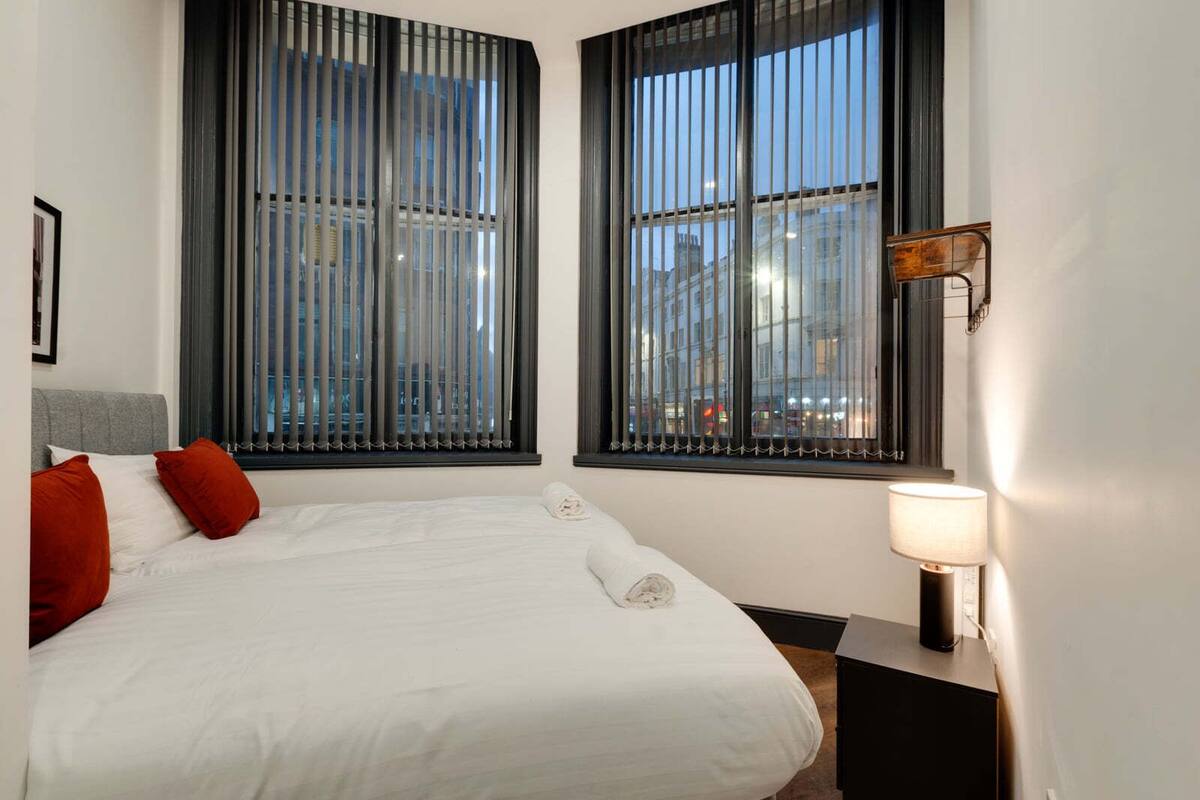 A hotel room with a large bed, two windows with blinds, a bedside lamp, and a view of the cityscape.