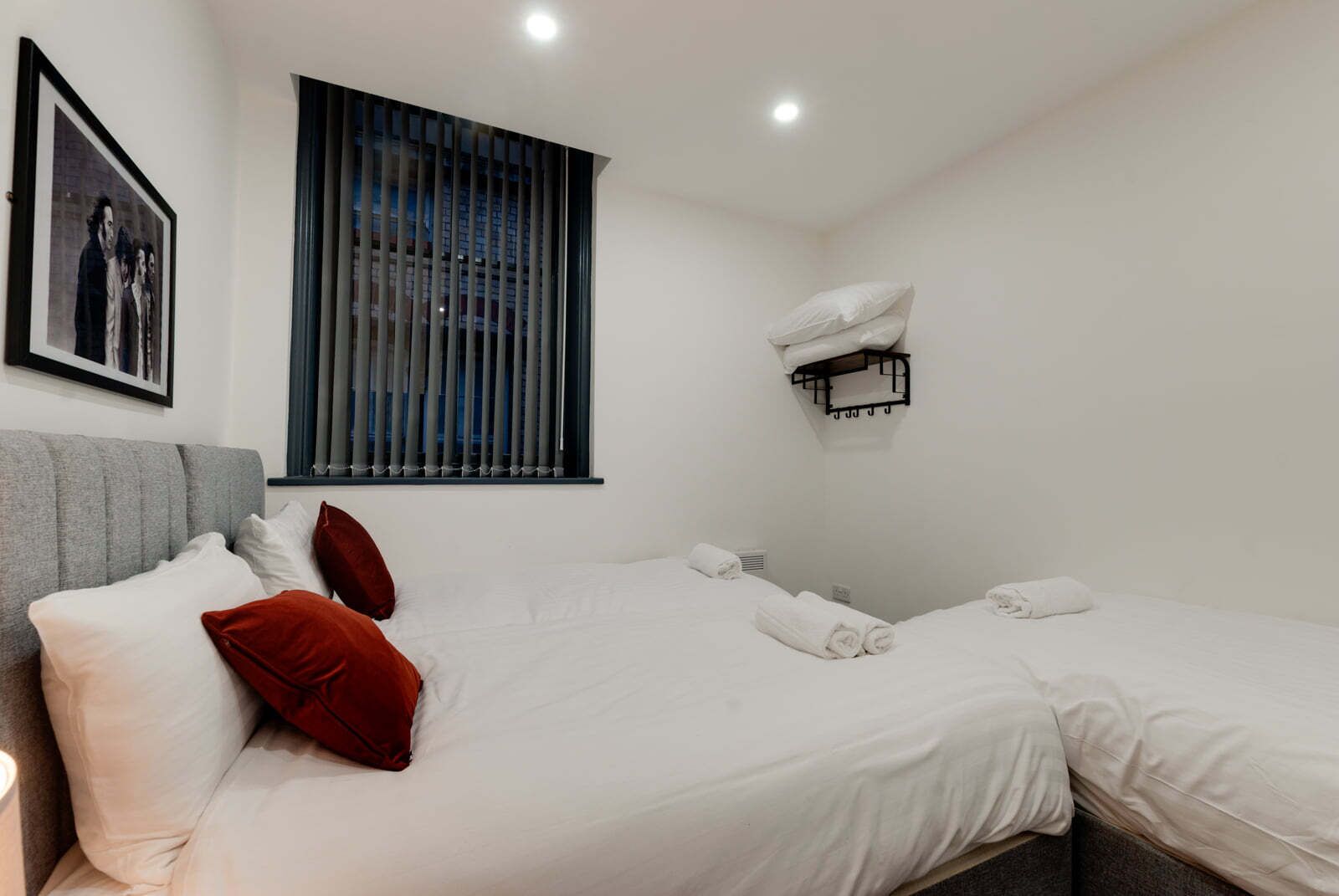 Une chambre d’hôtel avec deux lits, un canapé gris, un tableau encadré au mur, une fenêtre avec des stores et un porte-serviettes.