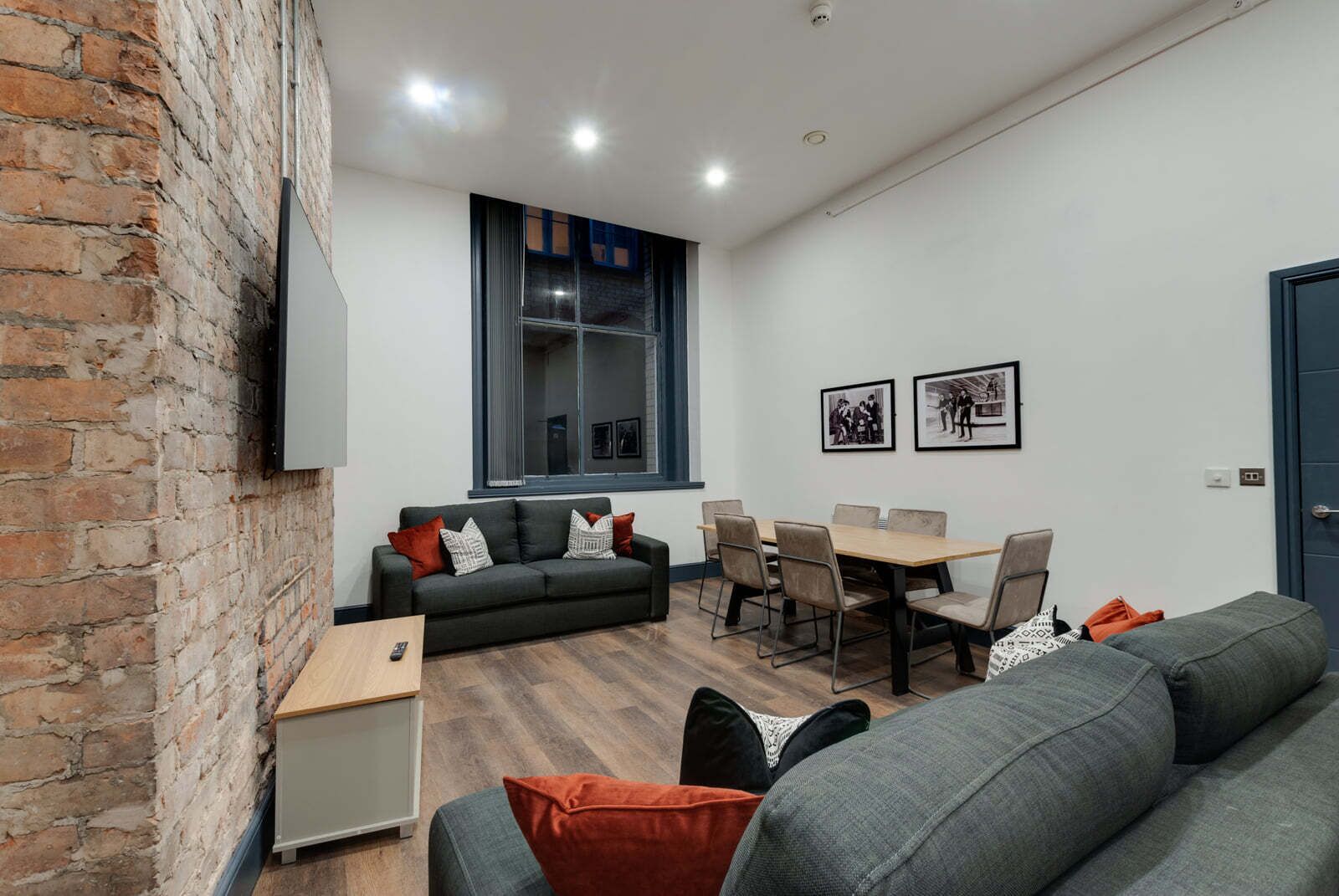 A modern living room with a brick wall, a grey sofa, a dining table with chairs, and a large window.
