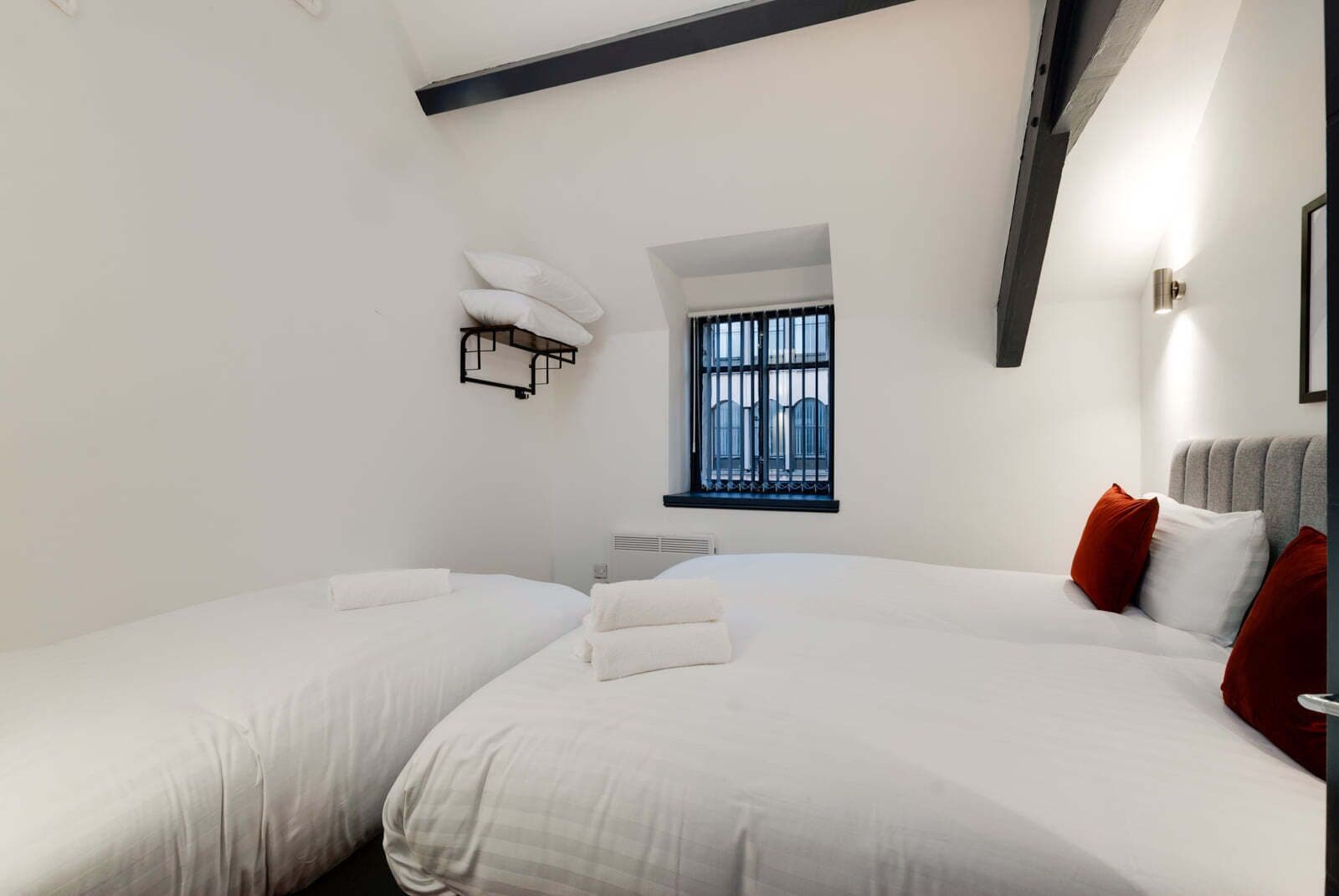 Habitación de hotel con dos camas, una ventana con barrotes, estante en la pared y mesita de noche.