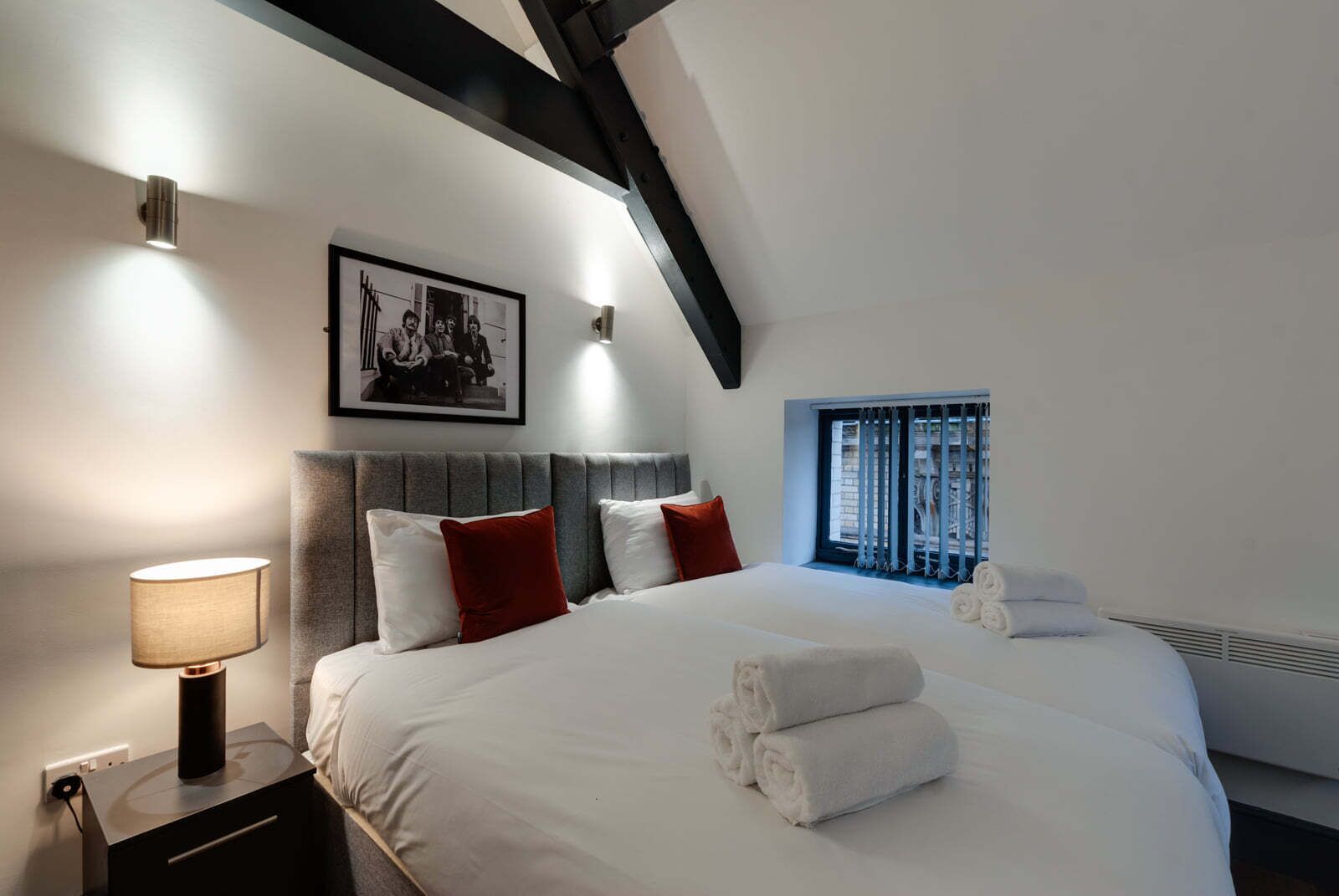 Un dormitorio con una cama grande, una mesita de noche con lámpara, un cuadro enmarcado en la pared y una ventana con persianas.