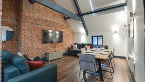 A modern living room with a brick wall, a flat-screen TV, a dining area with a table set for four, and a cozy seating area with a sofa and chairs. - Harrisons Aparthotel (Liverpool)